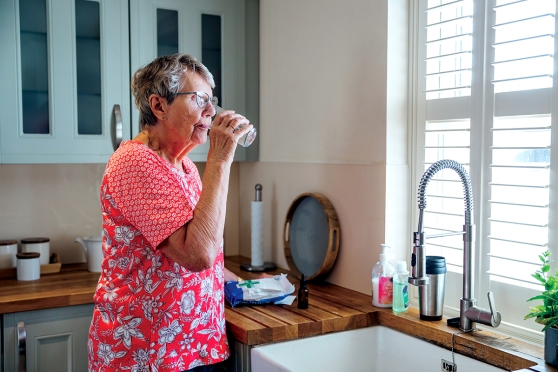 An older woman stands at her sink and drinks a glass of water.